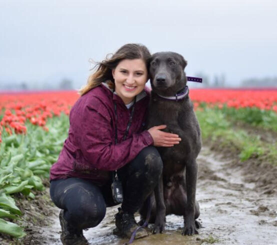 Trainer Ana Maria Thorne kneeling beside her dog Santos in a tulip field, smiling at the camera